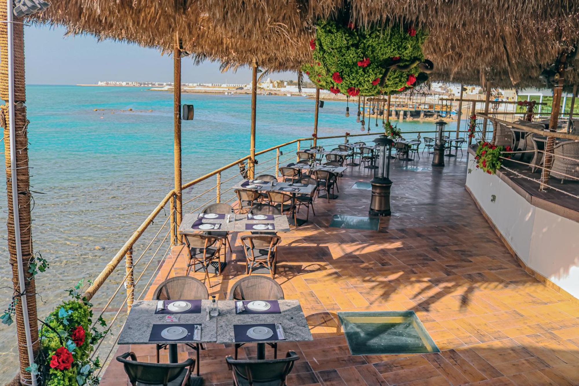 Lagoona Beach Luxury Resort And Spa Манама Екстериор снимка The photo shows a seaside restaurant with a spacious outdoor dining area. It features several tables set with plates and utensils, surrounded by lush greenery and decorative plants. The flooring is made of brown tiles, and there are thatched roofs pr
