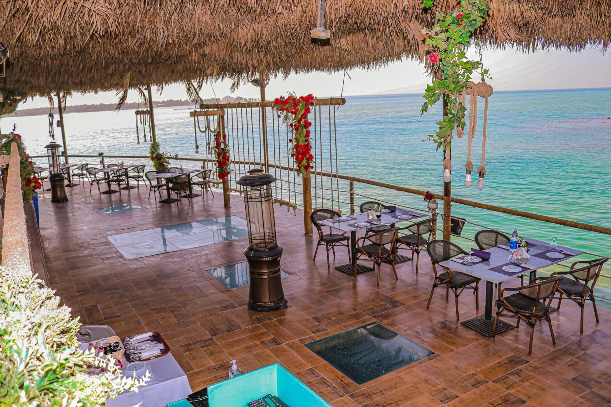 Lagoona Beach Luxury Resort And Spa Манама Екстериор снимка The photo depicts a coastal restaurant or dining area with an open-air design. It features a wooden floor and tables set for dining, surrounded by chairs. The space is partially covered by a thatched roof, providing shade while allowing for a view of