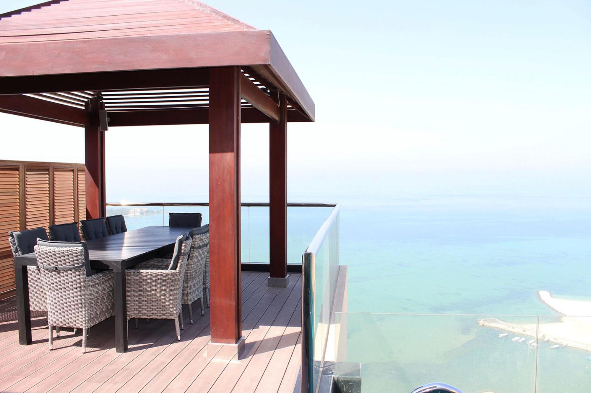 Lagoona Beach Luxury Resort And Spa Манама Екстериор снимка The photo showcases a stylish outdoor patio or terrace area. It features a wooden structure with a sloped roof, providing a shaded space. There is a large table made of dark wood, surrounded by several wicker chairs. The patio is elevated and has gla