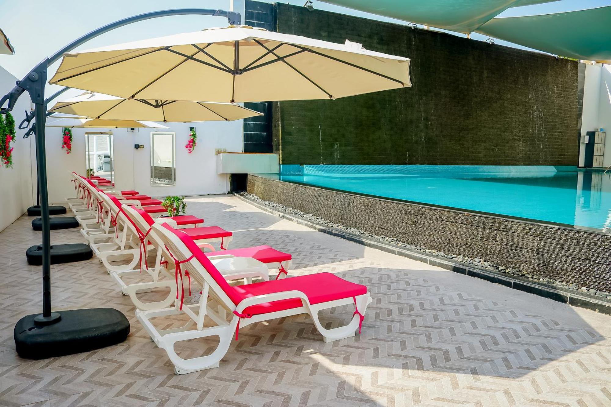 Lagoona Beach Luxury Resort And Spa Манама Екстериор снимка The photo shows a sunny poolside area with several lounge chairs arranged in a row. Each chair features red cushions and is positioned under large, beige umbrellas for shade. In the background, there is a beautiful swimming pool with clear blue water