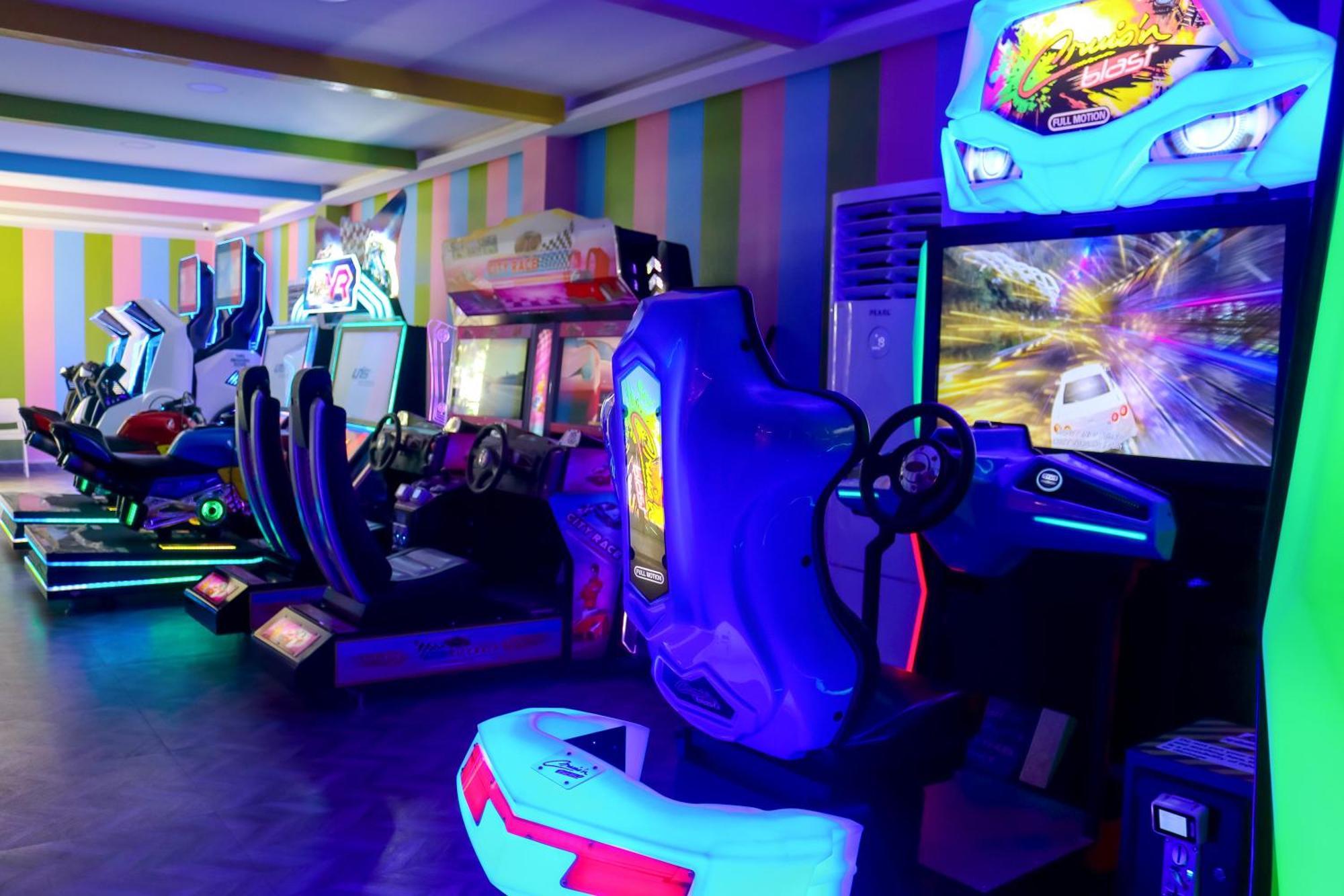 Lagoona Beach Luxury Resort And Spa Манама Екстериор снимка The photo shows an arcade area filled with various arcade machines. The setting is illuminated with colorful neon lights, creating a vibrant atmosphere. There are several racing game machines with steering wheels and seats, each accompanied by screen