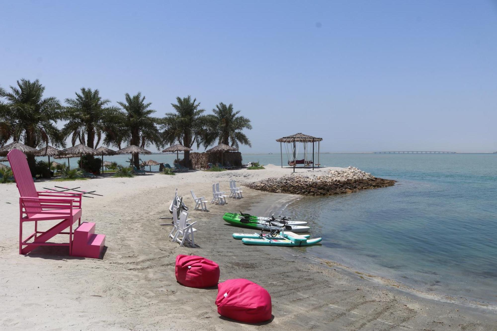 Lagoona Beach Luxury Resort And Spa Манама Екстериор снимка The photo depicts a serene beach scene with clear blue water and a sunny sky. In the foreground, there are colorful pink bean bags and a large pink chair on the sand. A few white chairs and loungers are positioned near the water's edge. To the side, 