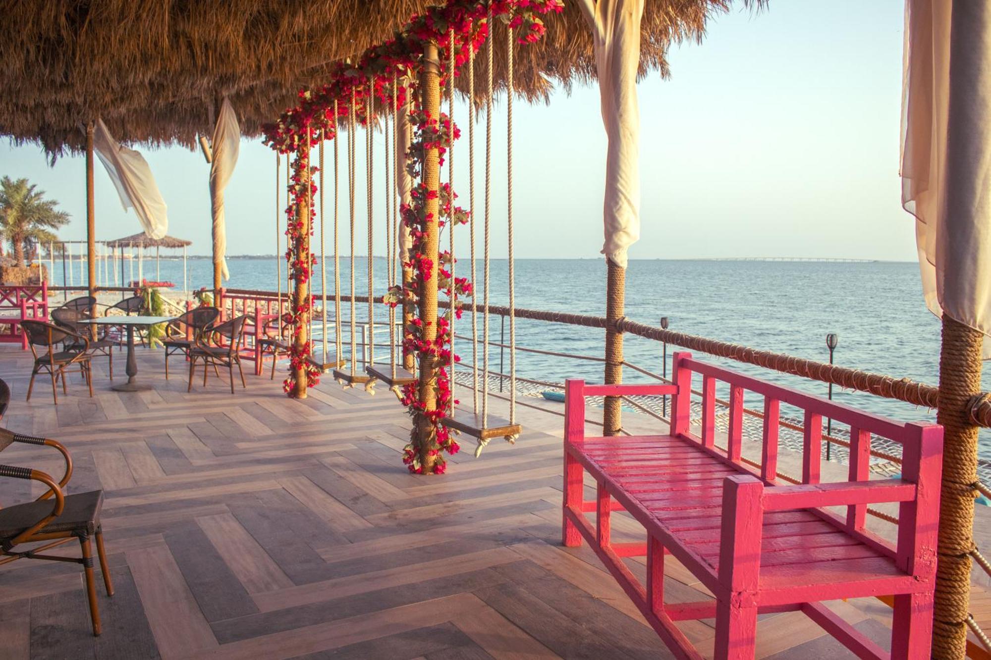 Lagoona Beach Luxury Resort And Spa Манама Екстериор снимка The photo depicts a scenic outdoor area by the water, with a wooden deck and a thatched roof. Two swings adorned with flowers are hanging from the ceiling, providing a charming touch to the space. In the foreground, there is a bright pink bench, addi