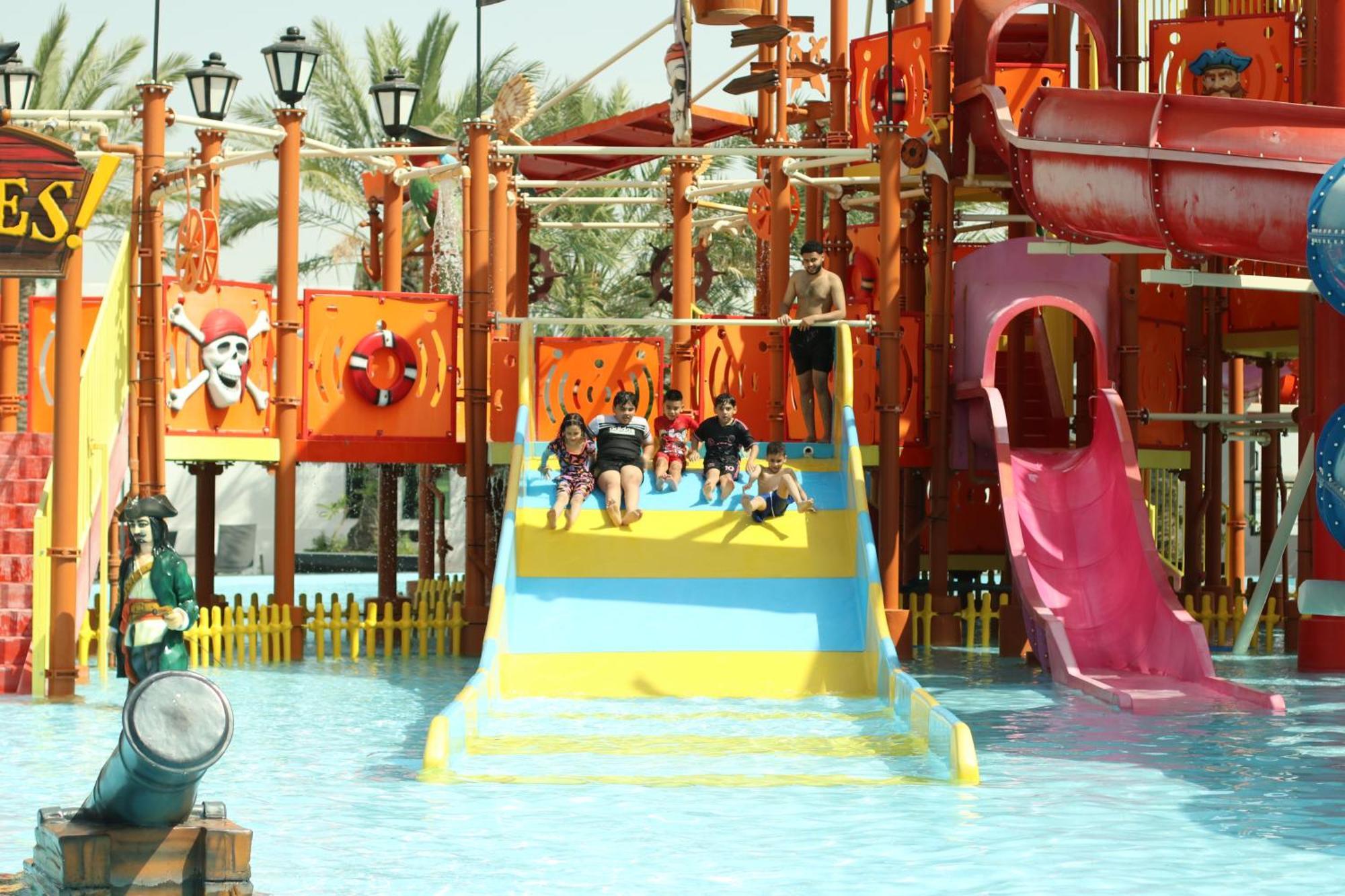 Lagoona Beach Luxury Resort And Spa Манама Екстериор снимка The photo shows a colorful water park scene featuring a play area with multiple slides. There are children enjoying themselves, sliding down bright yellow and pink slides into a pool of water. The structure has pirate-themed decorations, including a 