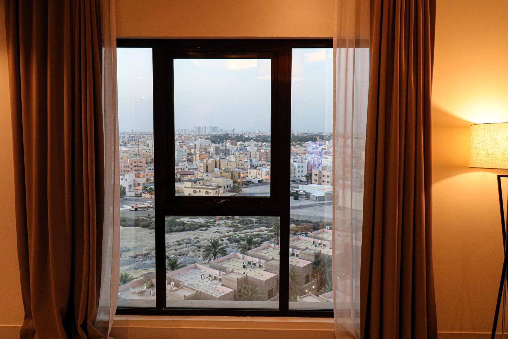 Lagoona Beach Luxury Resort And Spa Манама Екстериор снимка The photo shows a window with a black frame and sheer curtains. Through the window, there is a view of a cityscape, displaying a densely populated area with various buildings and rooftops. The sky appears slightly overcast, suggesting a muted lightin