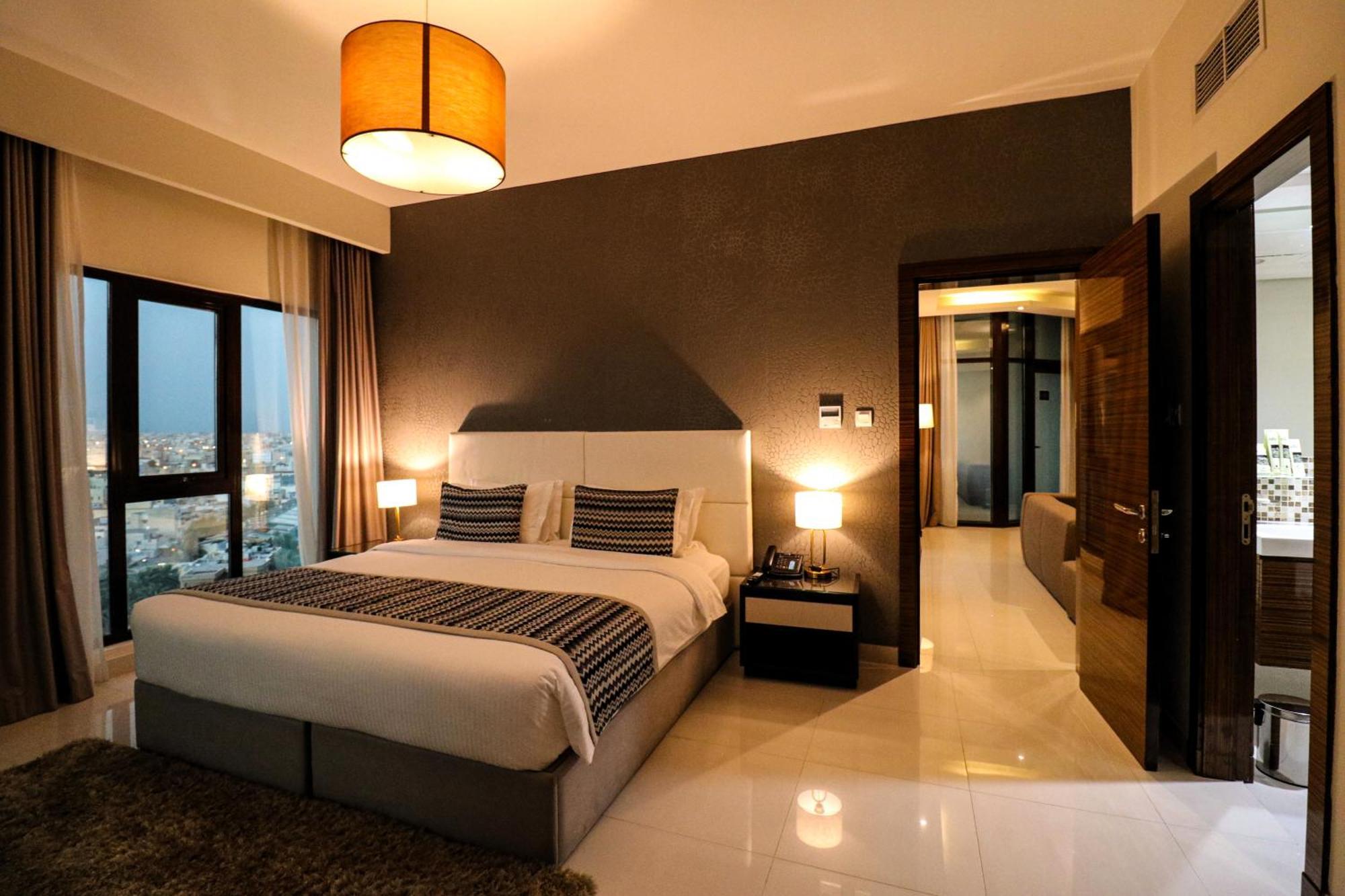 Lagoona Beach Luxury Resort And Spa Манама Екстериор снимка The photo depicts a modern hotel room or suite. It features a large bed with a white and gray color scheme, accented with decorative pillows. On either side of the bed, there are sleek bedside lamps providing warm lighting. The walls are a dark color