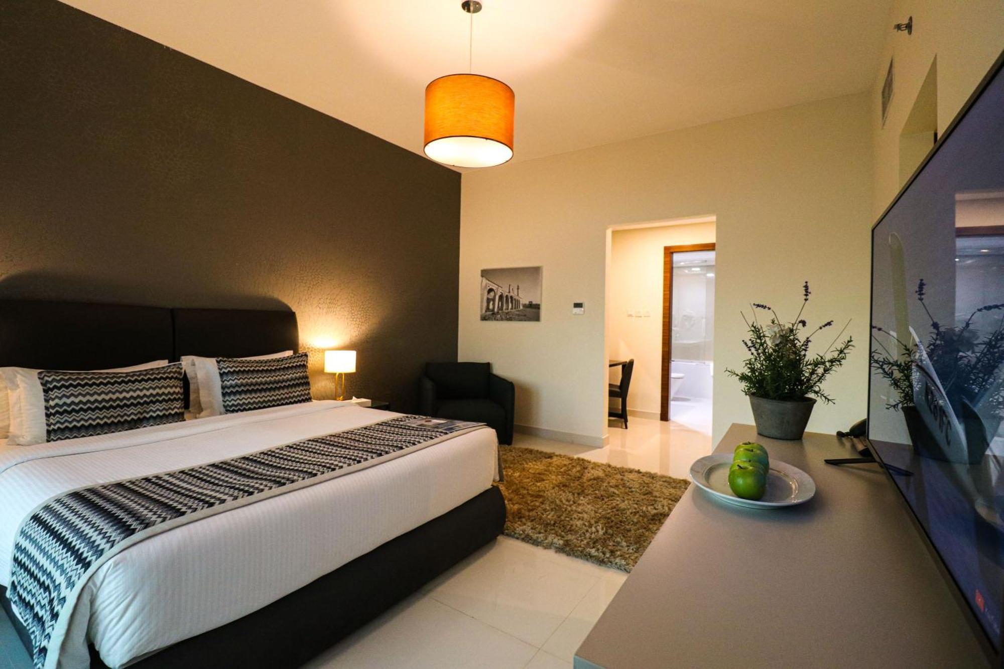 Lagoona Beach Luxury Resort And Spa Манама Екстериор снимка The photo shows a modern hotel room. It features a large bed with a neatly made white and dark-colored duvet. On either side of the bed, there are bedside tables with lamps. The wall behind the bed is painted in a dark color, creating a contrast with