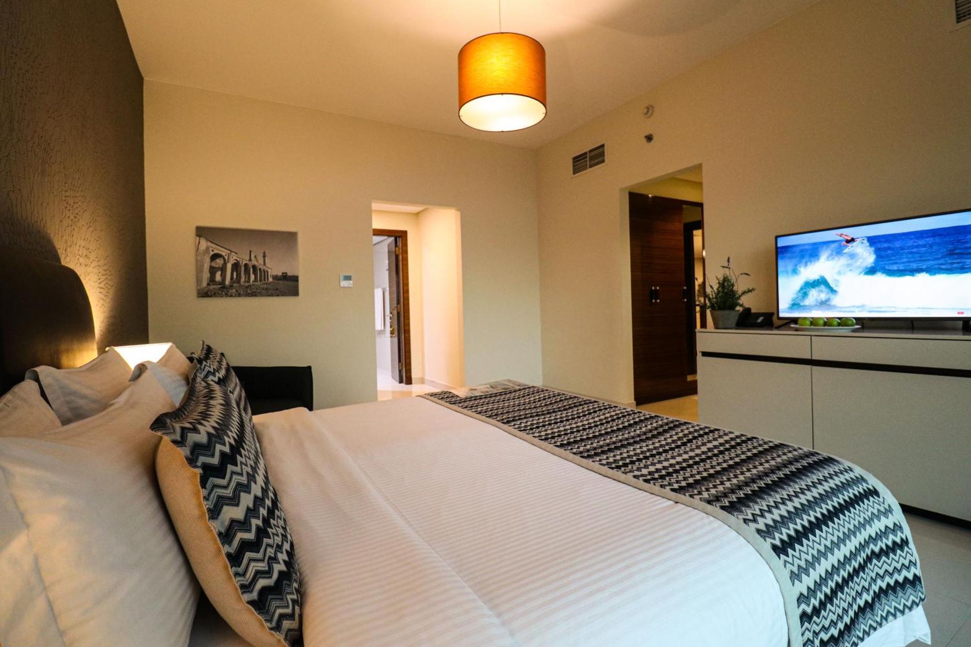 Lagoona Beach Luxury Resort And Spa Манама Екстериор снимка The photo shows a well-decorated hotel room. There is a large bed with white linens and decorative pillows featuring a zigzag pattern. On the wall beside the bed is a piece of artwork, and there is a modern television on a white furniture unit opposi