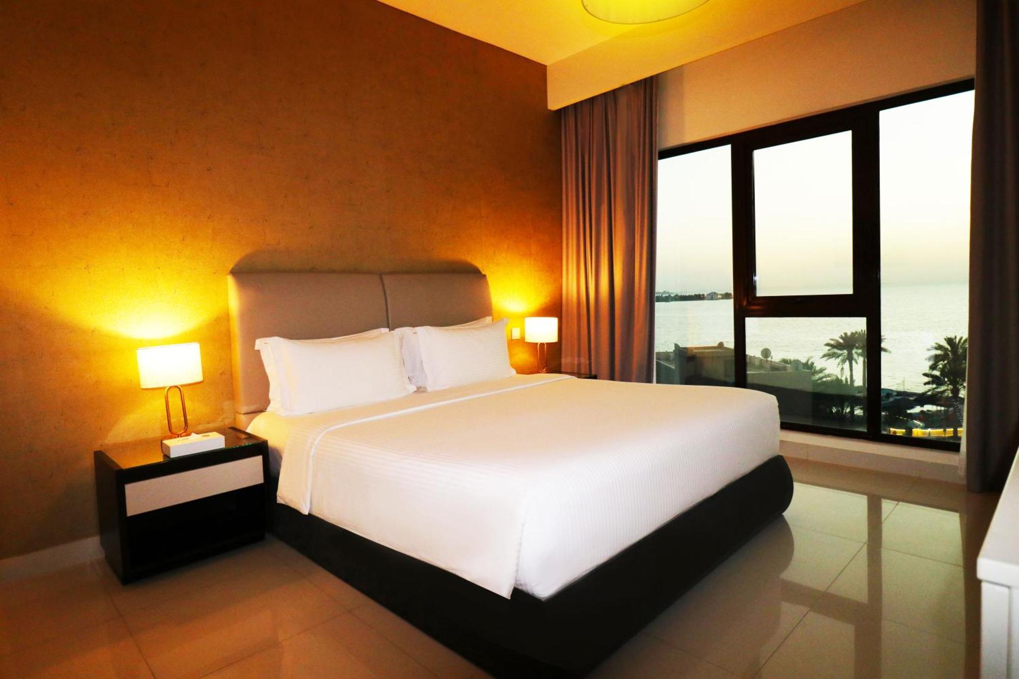 Lagoona Beach Luxury Resort And Spa Манама Екстериор снимка The photo shows a modern hotel bedroom with a large, comfortable bed dressed in white linens. The room features warm lighting, including bedside lamps, creating a cozy atmosphere. There are large windows with a view of the outdoors, possibly overlook