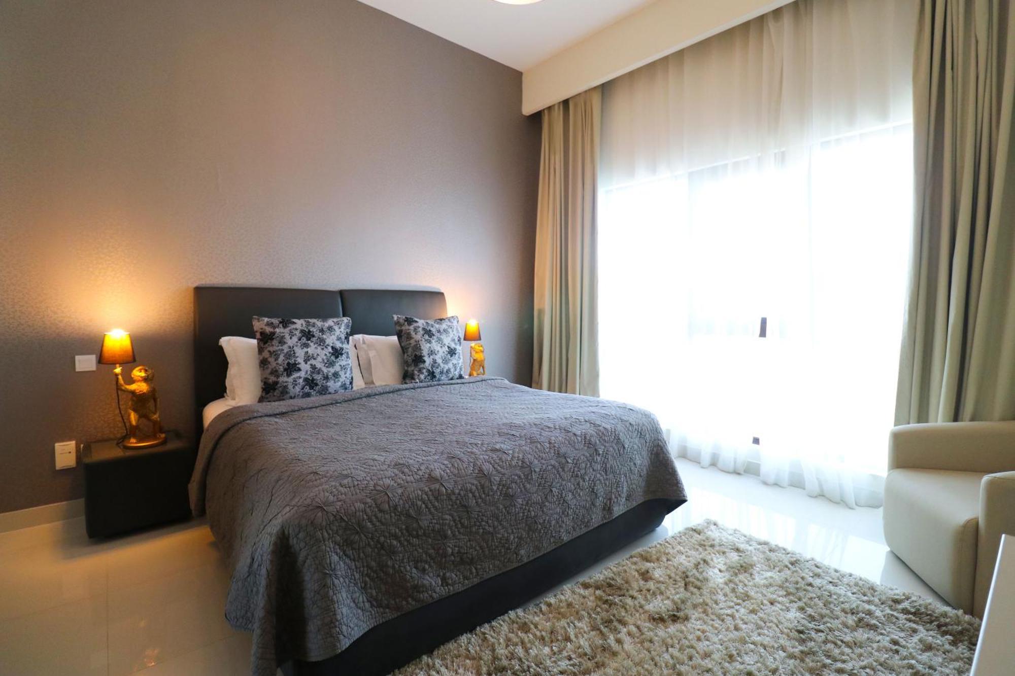 Lagoona Beach Luxury Resort And Spa Манама Екстериор снимка The photo shows a stylish bedroom with a modern design. It features a large bed with a dark gray bedspread and decorative pillows. On either side of the bed are two wooden nightstands, each with a lamp. There is a large window covered with light, flo